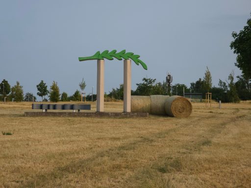 Grüne Skulptur auf Säulen, neben Rundballen in einem trockenen Feld.